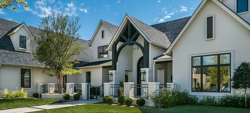 Modern suburban house with light-colored exterior, dark roof, large windows, front lawn, landscaped bushes, and a central arched entrance.