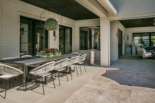 Covered outdoor patio with a long dining table, eight white chairs, a hanging round planter, and a heater; adjacent to an open lounge area.