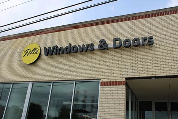 A brick building with large windows displays a “Pella Windows & Doors” sign in black letters next to a yellow logo.