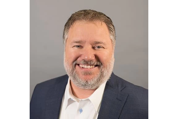 A middle-aged man with short brown hair and a gray beard, wearing a navy suit jacket and a white shirt, smiling in front of a plain gray background.