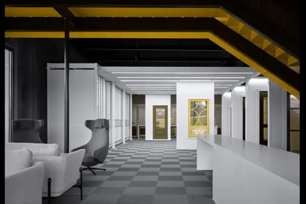 Modern office interior with checkered flooring, white walls, grey seating, large windows, and yellow structural beams.