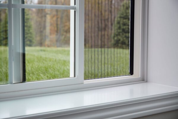 A close-up of a white-framed window partially open, with a mesh insect screen visible, overlooking green grass and trees outside.