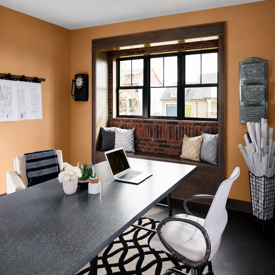 Modern home office with orange walls, a desk with a laptop and flowers, a window seat with pillows, wall drawings, and a basket of rolled papers next to a black rotary phone.